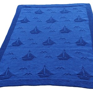 Può includere: Una coperta blu lavorata a maglia con un motivo di barche a vela e onde. La coperta ha un aspetto strutturato e un bordo dettagliato. Il tema nautico è enfatizzato dal ripetuto disegno della barca a vela.