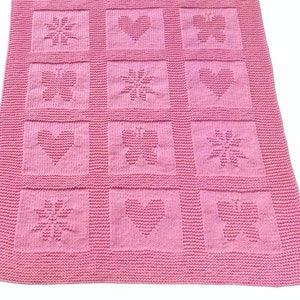 Può includere: Una coperta lavorata a maglia di colore rosa con una griglia di dodici pannelli quadrati. Ogni pannello mostra un motivo testurizzato a forma di cuore, farfalla o fiore simile a una stella. L'intero pezzo è realizzato con un punto uniforme.