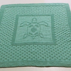 May include: A green knitted baby blanket with a turtle design in the center. The blanket is made with a textured stitch and has a border around the edges.
