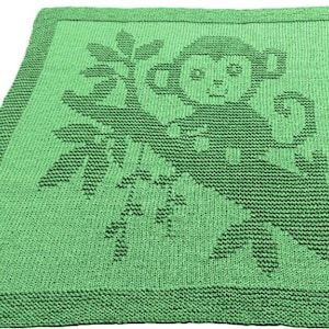 May include: A green knitted blanket featuring a monkey design. The monkey is depicted sitting on a branch with leaves. The blanket has a solid green border and is made of a soft, textured material.