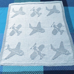 Könnte beinhalten: Eine weiße gestrickte Babydecke mit einem sich wiederholenden Muster aus Flugzeugen. Die Decke ist aus weichem Garn gefertigt und eignet sich perfekt als Geschenk zur Babyparty.