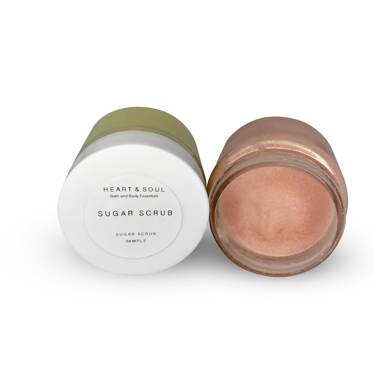 May include: Two open jars of sugar scrub. One jar is open, revealing a pinkish scrub. The other jar is closed with a white lid and a label that reads "HEART & SOUL Bath and Body Essentials SUGAR SCRUB SAMPLE."