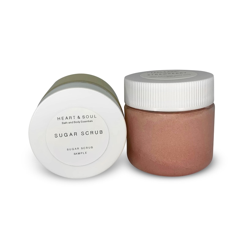 May include: Two jars of sugar scrub. One jar is lying on its side, displaying a white label with the text "HEART & SOUL" and "SUGAR SCRUB". The other jar is upright, filled with pink scrub, and has a white lid. The jars are for bath and body essentials.