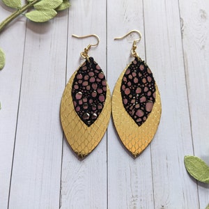 May include: A pair of gold and black dangle earrings with a pink and black patterned design. The earrings are made of leather and have a leaf shape.