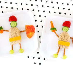 May include: Two white plates with a fun and healthy lunch idea for kids. Each plate features a sandwich, pretzel sticks, grapes, apple slices, cucumber, and carrot. The food is arranged to look like a knight.