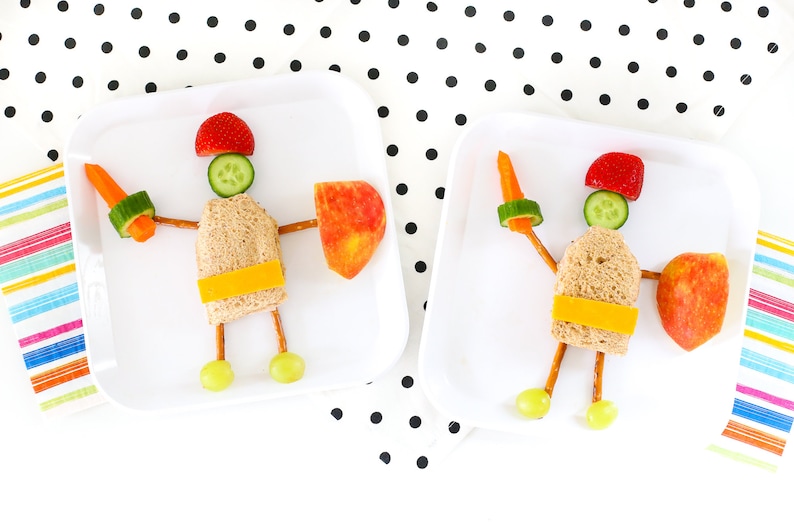 May include: Two white plates with a fun and healthy lunch idea for kids. Each plate features a sandwich, pretzel sticks, grapes, apple slices, cucumber, and carrot. The food is arranged to look like a knight.