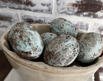 May include: A ceramic crock filled with blue and brown speckled Easter eggs. The crock is white with a rough texture and is sitting on a white surface.