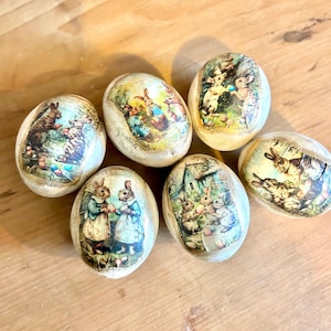May include: Six decorative Easter eggs with vintage-style rabbit illustrations. The eggs have a cream-colored base and are adorned with decoupage images of rabbits in various scenes, featuring pastel colors and a rustic aesthetic.
