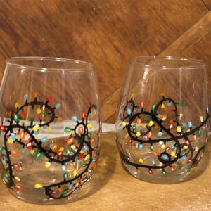 May include: Two clear glass wine glasses with hand-painted colorful Christmas lights. The lights are black outlined and have a variety of colors including red, yellow, blue, green, and white.