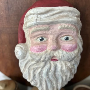 May include: A vintage-style Santa Claus face ornament. The ornament features a red hat, a white beard with glitter accents, and rosy cheeks. The eyes are blue, and the face is painted with a textured finish. A loop is attached for hanging.