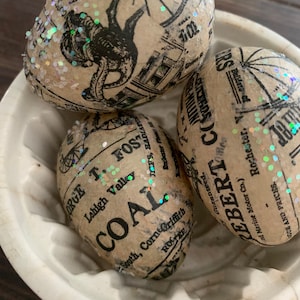 May include: Three decorative Easter eggs covered in brown paper with black text and white glitter. The eggs are decorated with vintage newspaper clippings and advertisements. The text on the eggs includes "Lehigh Valley", "Coal", "George T. Fost", "Guaranteed", "Rebert Co", "Rochester", and "Marine Motor Co."