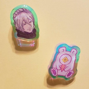 May include: Two iridescent resin pins. One features a character with white hair and a black scarf. The other features a pink creature with a single eye and a heart.