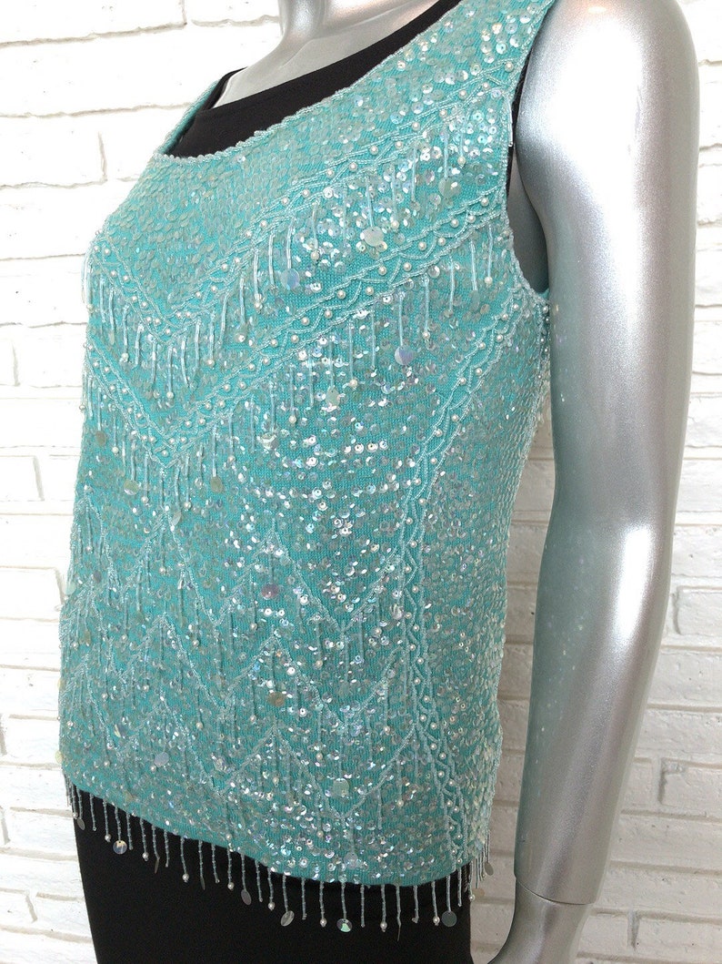 May include: A sleeveless turquoise sequined top with a beaded fringe trim. The top is made of a sheer fabric and has a scoop neckline.