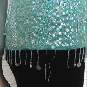 May include: A turquoise sequined sweater with a beaded fringe. The sweater has a delicate pattern of white beads and sequins.