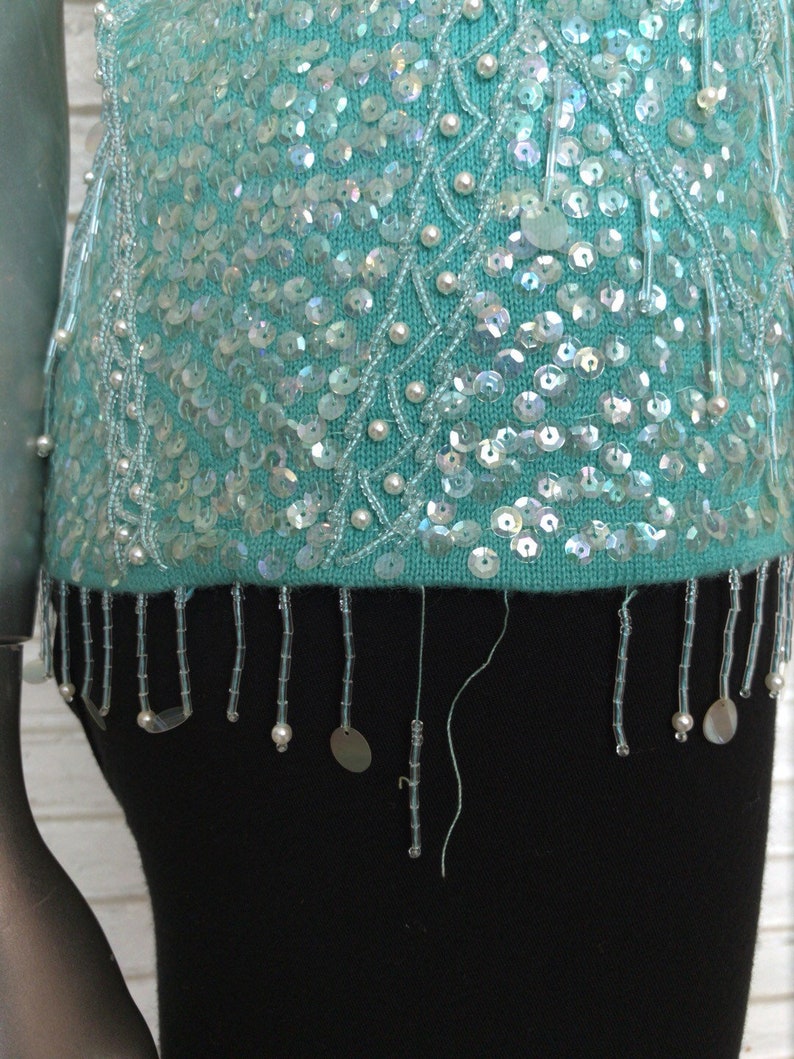 May include: A turquoise sequined sweater with a beaded fringe. The sweater has a delicate pattern of white beads and sequins.