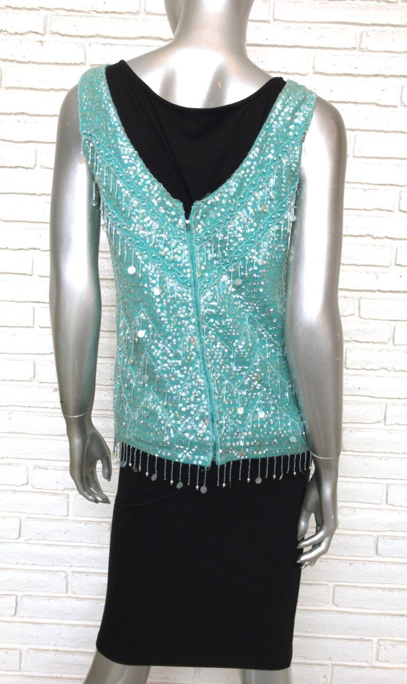 May include: A sleeveless turquoise sequined vest with a zipper down the back and a fringe trim. The vest is worn over a black top.