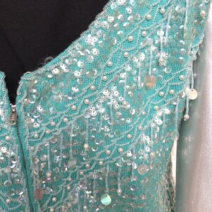May include: A teal blue knitted cardigan with a beaded design. The cardigan has a zipper closure and is embellished with iridescent sequins and white beads.