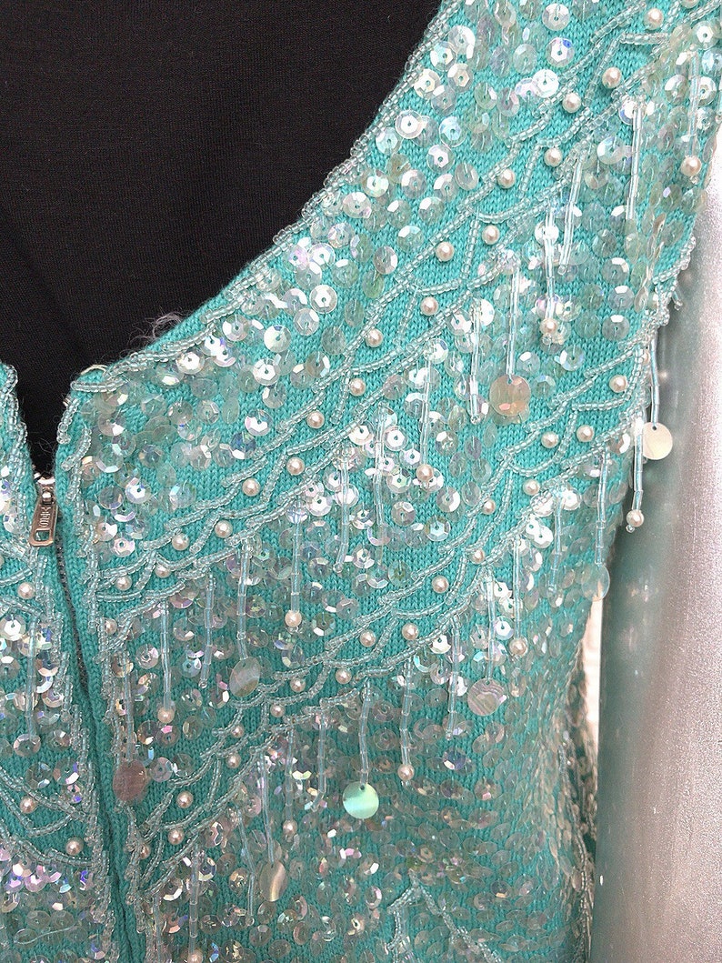 May include: A teal blue knitted cardigan with a beaded design. The cardigan has a zipper closure and is embellished with iridescent sequins and white beads.