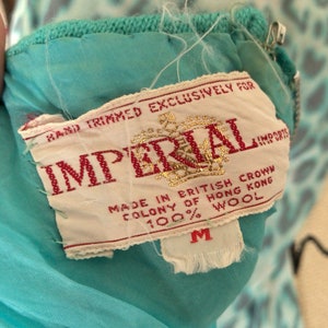 May include: A white tag with red text that reads "Hand Trimmed Exclusively For Imperial Imports" and "Made in British Crown Colony of Hong Kong 100% Wool". The tag is attached to a turquoise garment with a zipper.