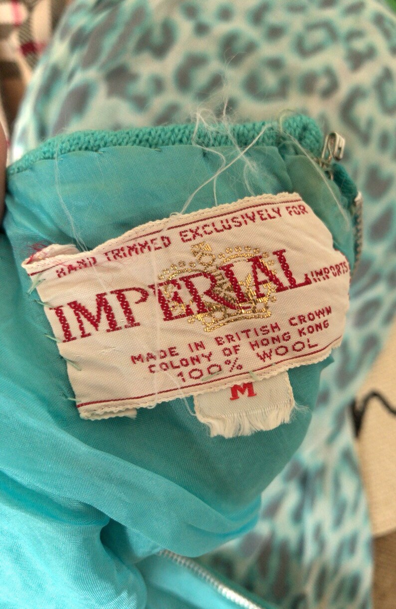 May include: A white tag with red text that reads "Hand Trimmed Exclusively For Imperial Imports" and "Made in British Crown Colony of Hong Kong 100% Wool". The tag is attached to a turquoise garment with a zipper.