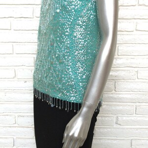 May include: A turquoise sequined sleeveless top with a beaded fringe trim. The top is on a white brick background.