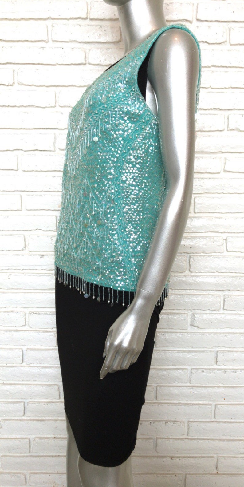 May include: A turquoise sequined sleeveless top with a beaded fringe trim. The top is on a white brick background.