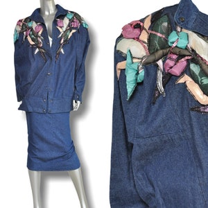 May include: A denim jacket and skirt set with colorful floral embellishments. The jacket has a button-up front and long sleeves. The skirt is a pencil skirt.