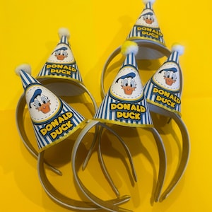 May include: A collection of Donald Duck party hats on headbands. The hats are blue and white striped with a yellow border and feature Donald Duck's face and the words "DONALD DUCK". Each hat is topped with a white pom-pom.