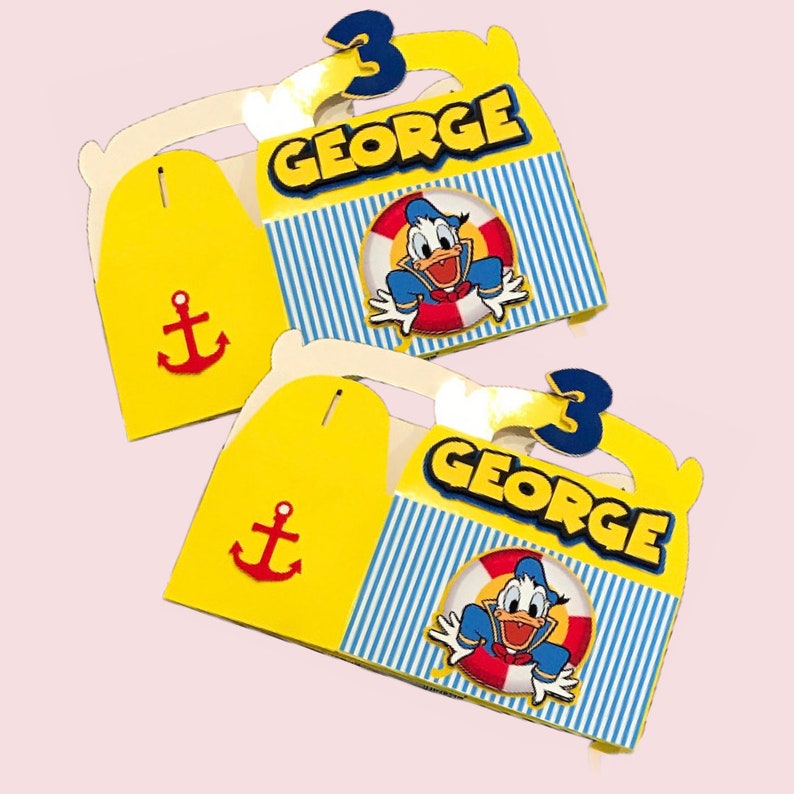 Pode incluir: Duas caixas de lembrancinhas amarelas e azuis com uma imagem de desenho animado do Pato Donald usando um salva-vidas. As caixas t&ecirc;m um padr&atilde;o listrado azul e branco e dizem "GEORGE" e "3" nelas. Cada caixa tem uma &acirc;ncora vermelha na frente.