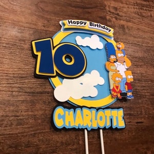 May include: A blue and yellow cake topper with the Simpsons family, the number 10, and the name Charlotte. The topper says "Happy Birthday" on a yellow banner.