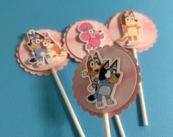 May include: Four pink and white cupcake toppers with different characters from the cartoon show Bluey. The characters are Bluey, Bingo, Bandit, and Chilli. The toppers are on white sticks.