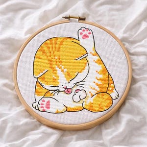 Könnte beinhalten: Ein Kreuzstich-Stickbild in einem runden Holzrahmen, das eine sich putzende orange-weiß getigerte Katze zeigt. Die Katze ist mit erhobenem Bein dargestellt, wobei rosa Pfotenballen und eine kleine rosa Zunge auf weißem Stoff zu sehen sind.