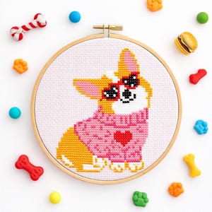 Op de afbeelding: Een kruissteekborduurwerk in een ronde houten borduurring met een geel-witte Corgi-hond die een roze trui met een rood hart draagt. De hond heeft zwarte hartvormige ogen. Kleine kleurrijke plastic hondenthema-bedels omringen de borduurring.