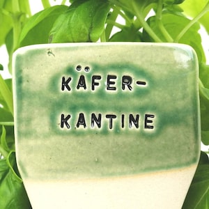 Könnte beinhalten: Ein rechteckiger, hellgrüner Keramik-Pflanzenmarker mit dem Text "KÄFER-KANTINE" in Schwarz. Der Marker hat eine weiße Basis und ist von grünen Basilikumblättern umgeben.