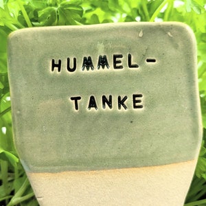 Könnte beinhalten: Ein rechteckiger, hellgrüner Keramik-Gartenmarker mit den Worten "HUMMEL-" und "TANKE" in schwarzer, geprägter Schrift. Der untere Rand ist hellbeige. Der Marker befindet sich vor einem Hintergrund aus grünem Laub.