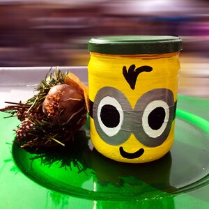 Op de afbeelding: Een gele glazen pot met een groen deksel, geschilderd om eruit te zien als een minion-personage. De pot heeft een zwart ooglapje, grote zwarte ogen en een glimlachende mond. De pot staat op een groen bord.