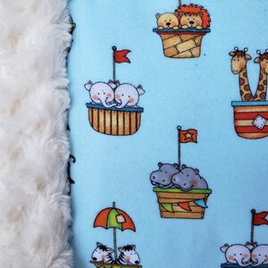 May include: A blue fabric with a white fluffy border. The fabric features a repeating pattern of animals riding in small boats. The animals include elephants, giraffes, hippos, monkeys, and lions. The boats are decorated with flags and stripes.