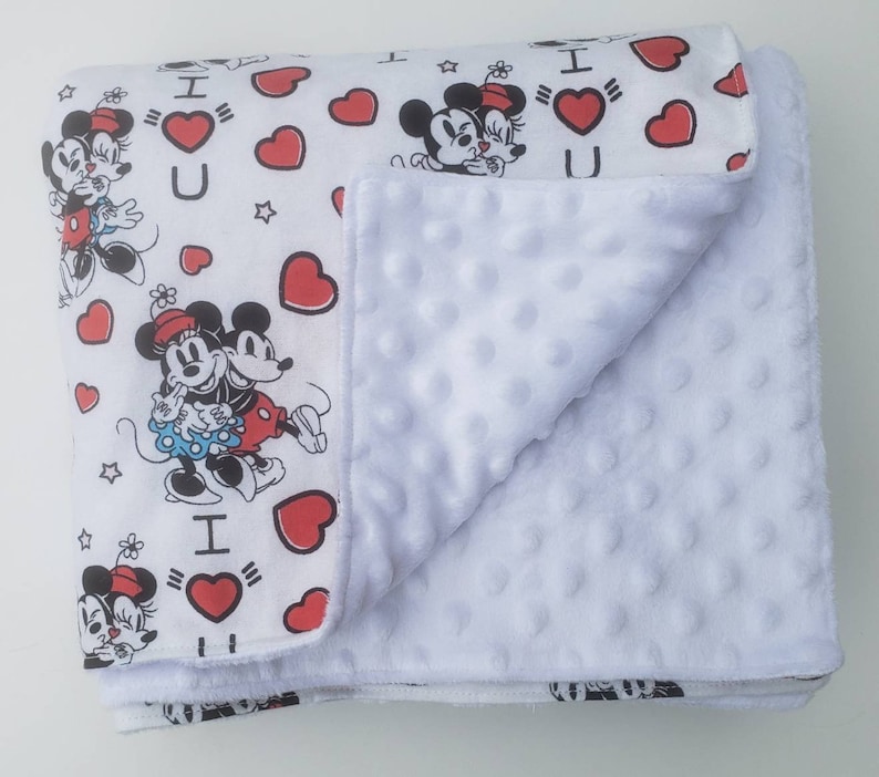 May include: White baby blanket featuring a pattern of Mickey and Minnie Mouse, red hearts, and the text "I ❤️ U." The blanket has a soft, textured white underside. The blanket is folded, showing both the patterned top and the plush underside.