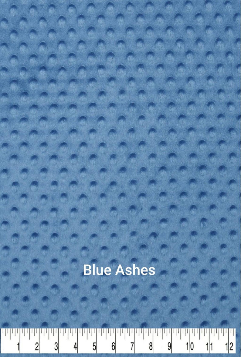 May include: Close-up of a blue fabric with a textured, dimpled surface. The fabric is a solid color, with the text "Blue Ashes" printed on it. A ruler is at the bottom, showing measurements in inches.