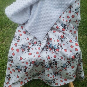 May include: A white baby blanket with a Mickey Mouse pattern and red hearts. The blanket has a soft, textured white backing. The blanket is draped over a wooden rocking chair.