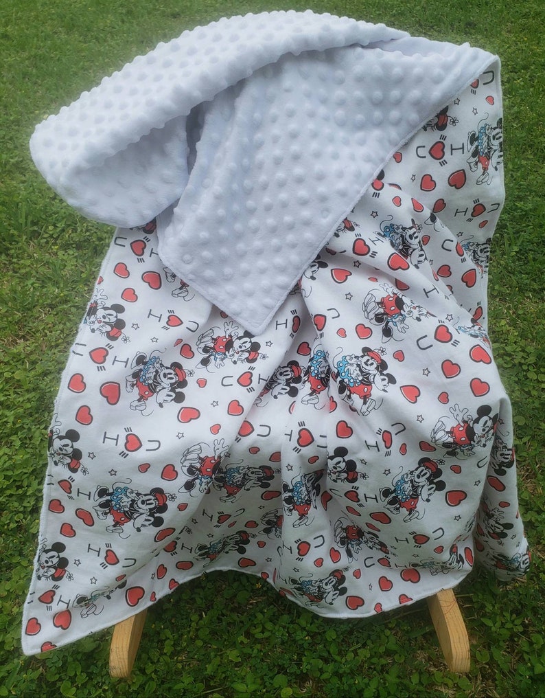 May include: A white baby blanket with a Mickey Mouse pattern and red hearts. The blanket has a soft, textured white backing. The blanket is draped over a wooden rocking chair.