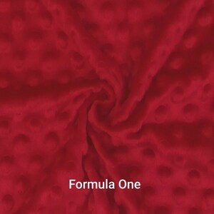 May include: A close-up of a red fabric with a textured, dotted pattern. The fabric is a deep, rich red color. The text "Formula One" is displayed in white at the bottom of the image. The fabric appears soft and plush.