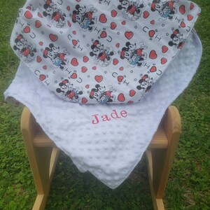 May include: A white baby blanket with a Mickey Mouse pattern and the name "Jade" embroidered in pink. The blanket is placed on a wooden rocking chair, with a green grass background.