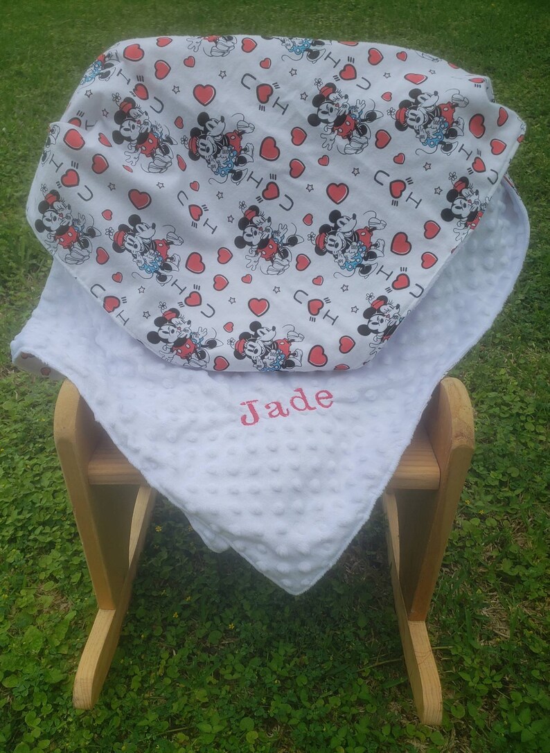 May include: A white baby blanket with a Mickey Mouse pattern and the name "Jade" embroidered in pink. The blanket is placed on a wooden rocking chair, with a green grass background.