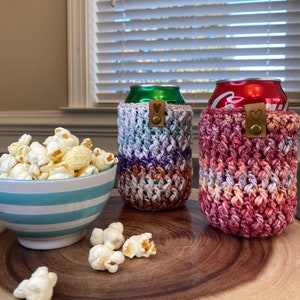 May include: Two crocheted can cozies in different colors. The cozy on the left is a mix of purple, blue, and white. The cozy on the right is a mix of red, orange, and white. Both cozies have a brown heart-shaped button.