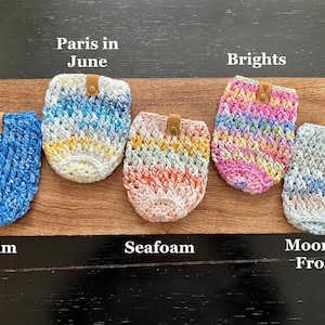 May include: Five crocheted cup cozies in different colors and patterns. The cozies are labeled "Denim", "Seafoam", "Moonlit Frost", "Paris in June", and "Brights".