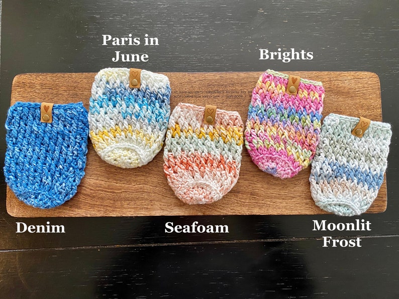 May include: Five crocheted cup cozies in different colors and patterns. The cozies are labeled "Denim", "Seafoam", "Moonlit Frost", "Paris in June", and "Brights".