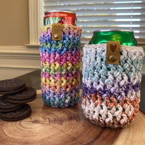 May include: Two crocheted can cozies, one in rainbow colors and one in shades of blue, purple, and brown. Both cozies have a brown leather strap with a heart-shaped metal accent.