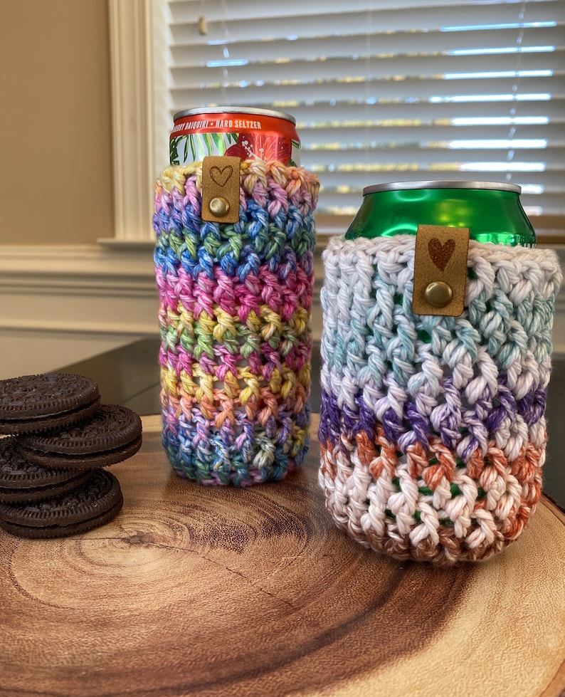 May include: Two crocheted can cozies, one in rainbow colors and one in shades of blue, purple, and brown. Both cozies have a brown leather strap with a heart-shaped metal accent.