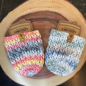 May include: Two crocheted can cozies in a rainbow and blue color scheme. The cozies are handmade and have a brown tag that says "handmade CAN COZY" with a heart.